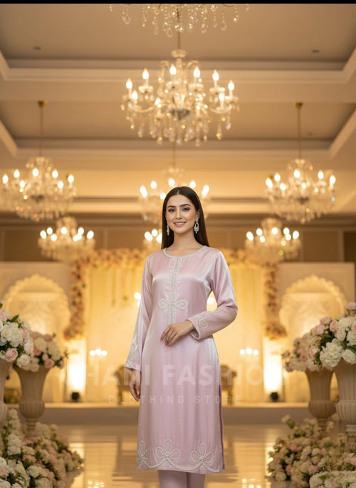 A woman stands in a grand, warmly lit event hall wearing a light pink traditional long-sleeve kurta dress with white embroidery. Crystal chandeliers and floral arrangements are visible in the background.