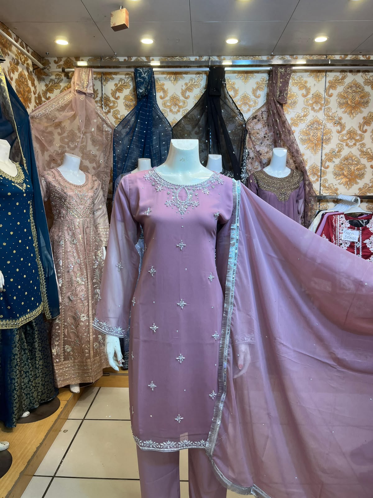 A lavender purple traditional salwar kameez suit displayed on a mannequin in a clothing store. The set includes a long-sleeved embroidered kameez top, matching trousers, and a sheer lavender dupatta with silver border detailing.