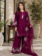 A woman models a deep magenta three-piece traditional outfit with silver mirror work embroidery, consisting of a long kurta, wide-leg trousers, and a matching dupatta, set against an elegant indoor garden patio backdrop.
